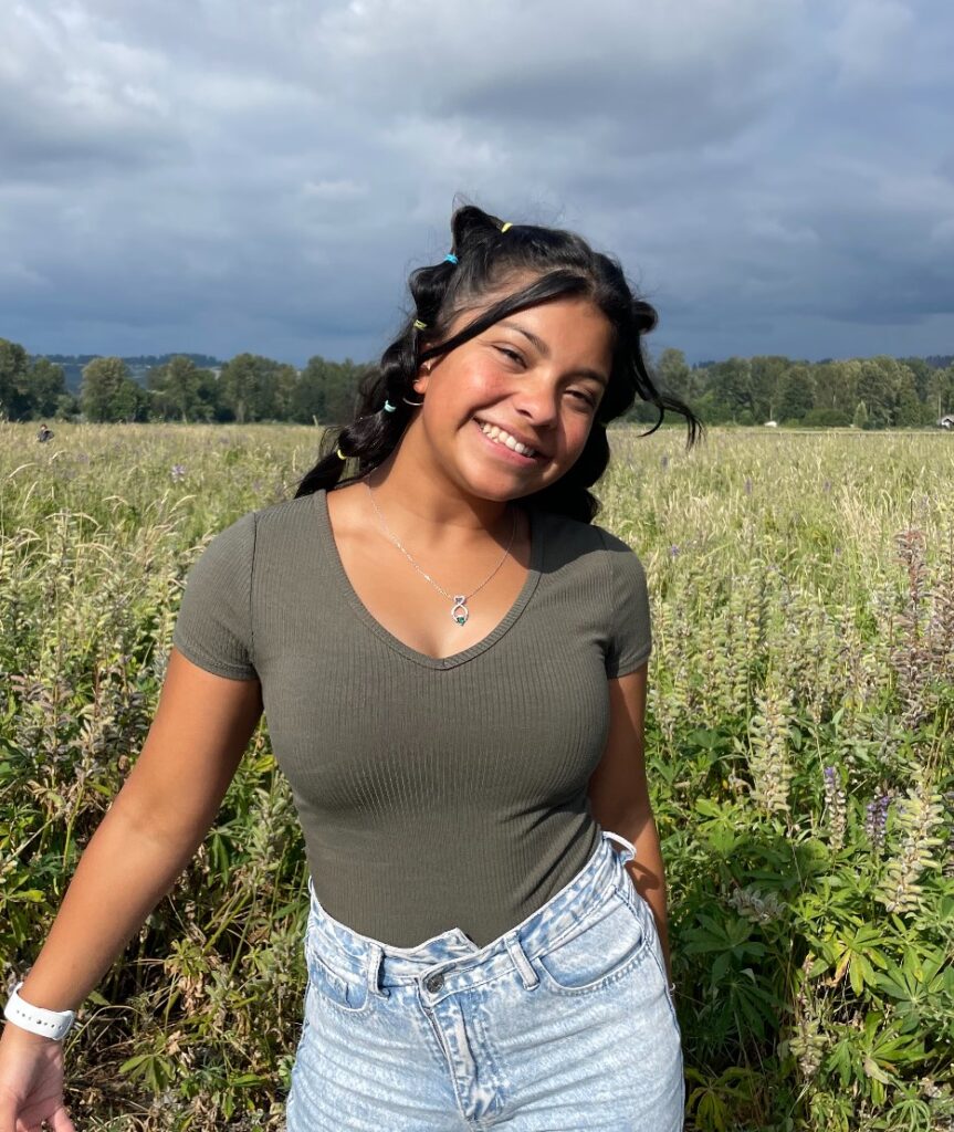 Cymi standing in front of a field, smiling with her arms outstretched.
